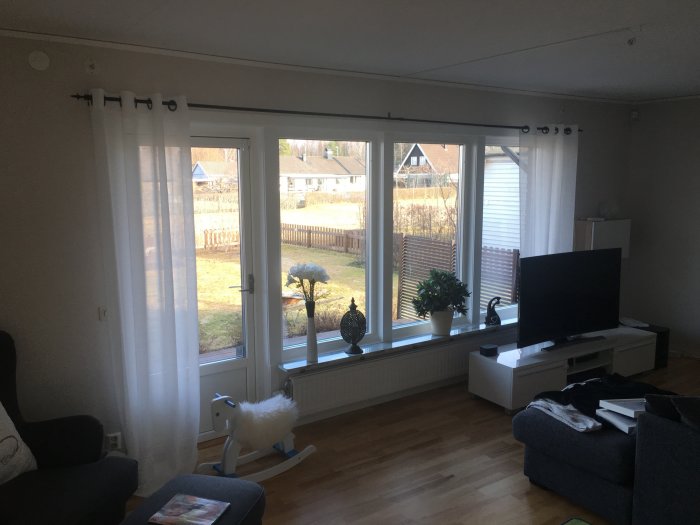 Vardagsrum med stor fönster som behöver rullgardin för att blockera morgonsol, tv och soffa syns.