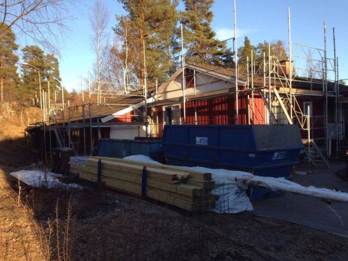 Byggarbetsplats med ställningar runt en bostad, virke och containers i trädgården.