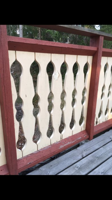 Delvis ruttet och flagnande rött verandastaket med ornamenterade vita paneler, visas framför grå trätrall och grönska.