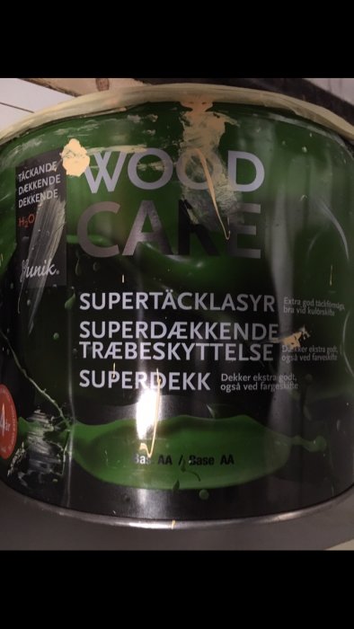 Burk med träskyddsfärg märkt "WOODCARE" och "SUPERTÄCKLASYR" med gröna stänk.