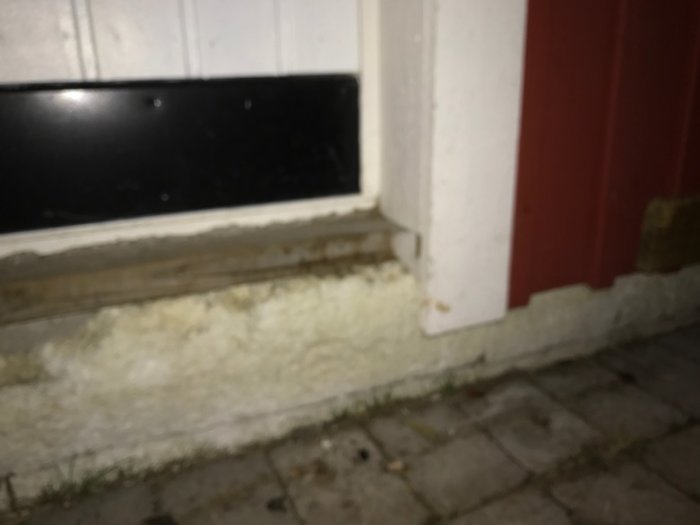 Synlig vittrande cellplast vid husgrund intill en dörr med svart bockad plåt och vit vägg ovanför.