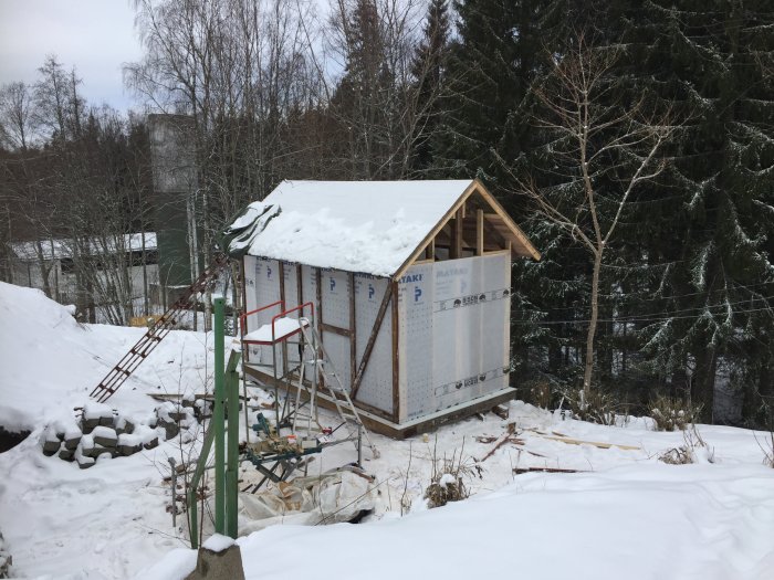 Stomme till en bastubyggnad i ett snöigt landskap med vindpapp och början till läkt, byggmaterial i förgrunden.