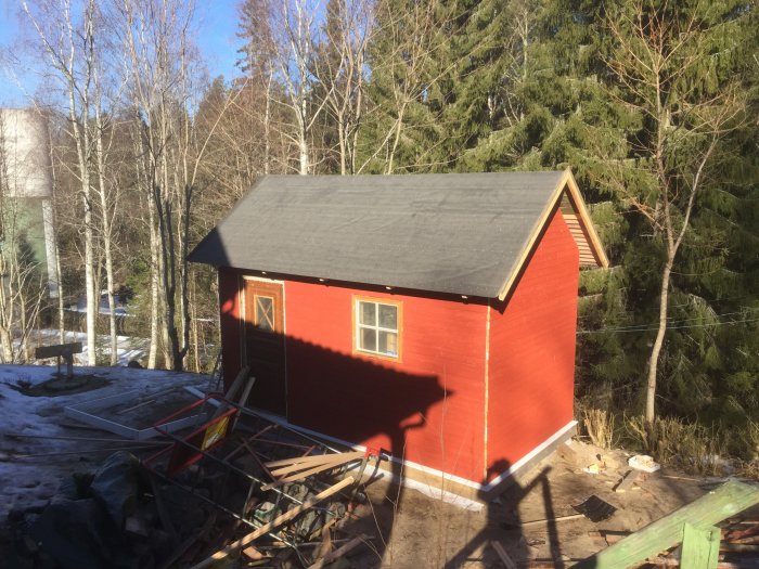 Rödmålad bastubyggnad under konstruktion med plåttak och trädetaljer, omgiven av snö och skog.