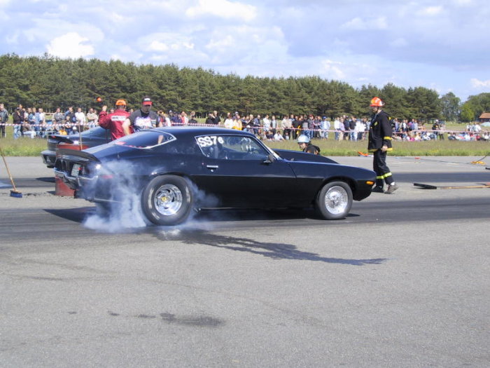 Svart muskelbil gör burnout vid dragracing evenemang med publik i bakgrunden.