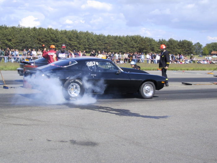 Svart muskelbil gör burnout på racingbana med publik i bakgrunden.
