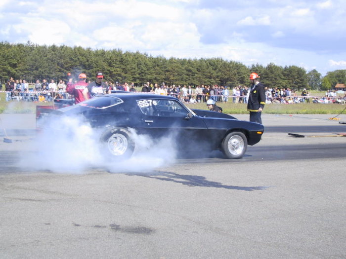 Svart bil gör burnout på en racingbana med publik i bakgrunden och säkerhetspersonal nära.