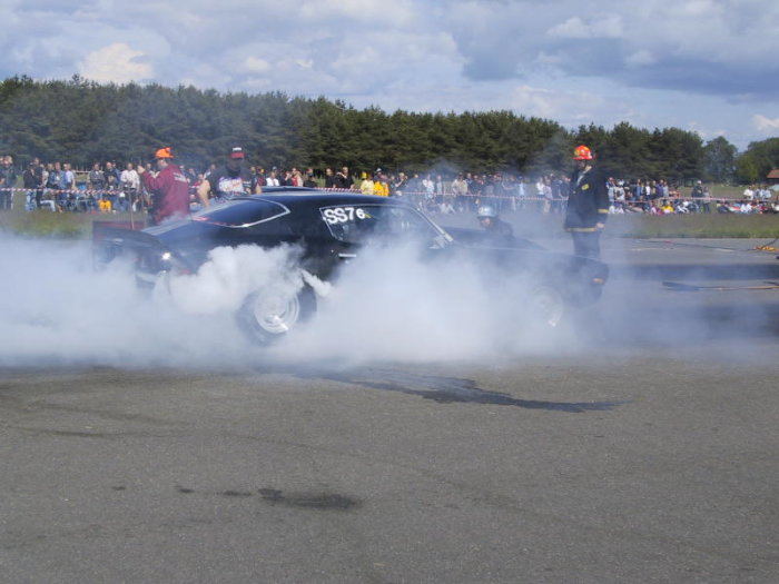 Svart bil utför burnout på tävling med däckrök och publik i bakgrunden.