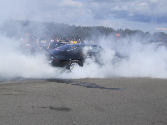 Svart bil gör en burnout och producerar mycket rök på en motorsportbana med publik i bakgrunden.