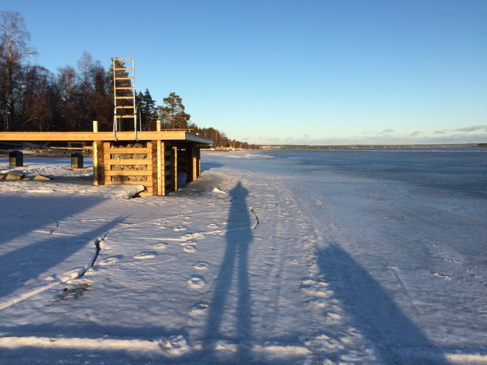 Brygga på vintertid med snö och istäckt vatten i bakgrunden, lång skugga i förgrunden.