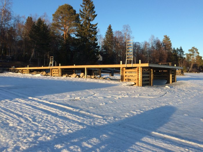 En brygga omgiven av snö med en stående stege, i ett vinterlandskap under solig himmel.