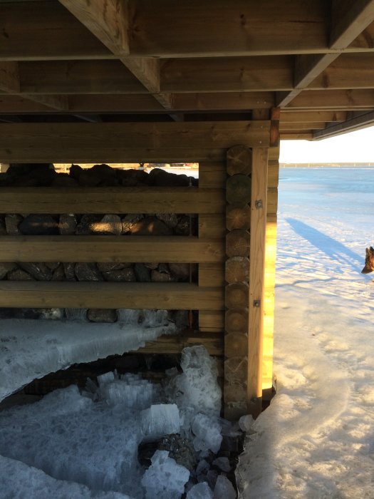 Träbrygga på strand med isbildningar under, mot solnedgång över frusen sjö.