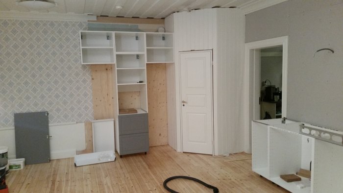 Delvis installerade vita köksstommar och skåp i ett pågående renoveringsprojekt med trägolv och en stängd dörr i bakgrunden.