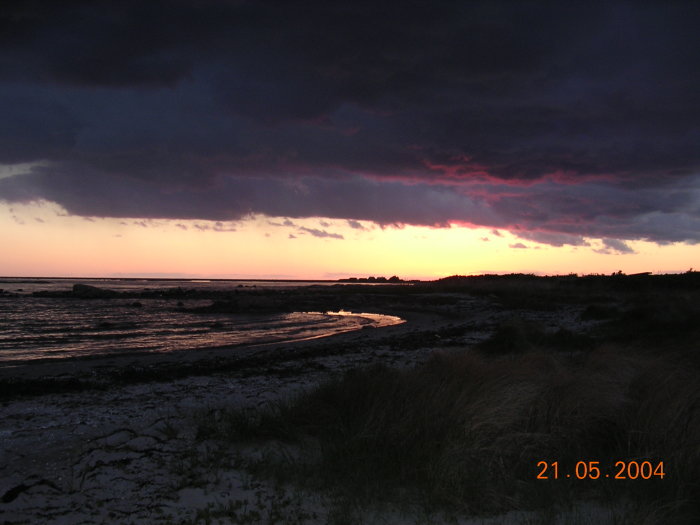 Solnedgång över Särdals strand med siluetter av fiskebodar och livräddningsstation mot dramatisk himmel.