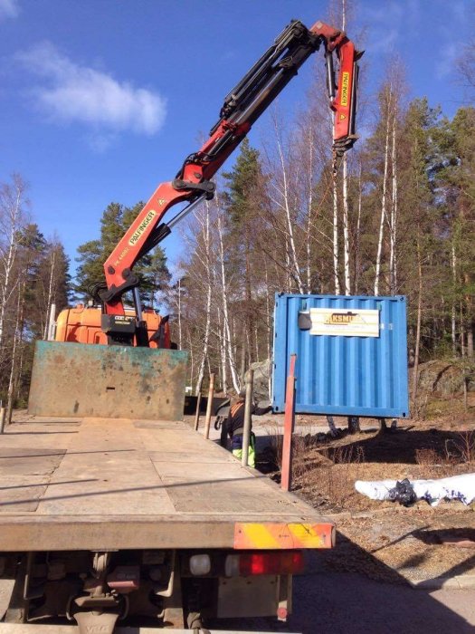 Kran som lyfter en blå verktygscontainer från en släpvagn mot en skogsbakgrund.