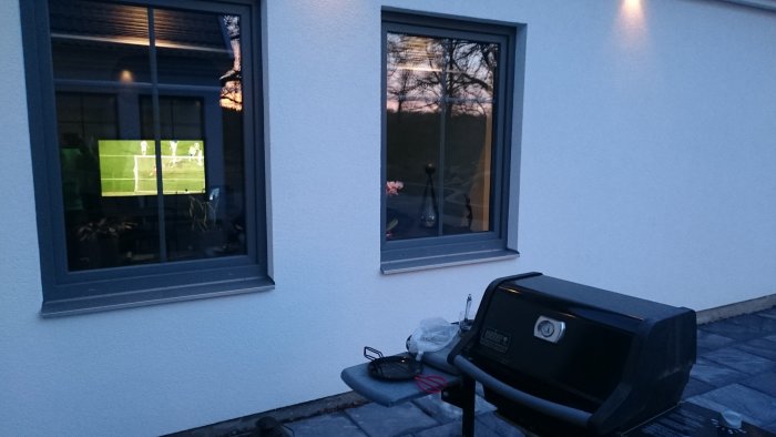 En äldre Weber-grill framför ett hus med fotbollsmatch i tv synlig genom fönstret.