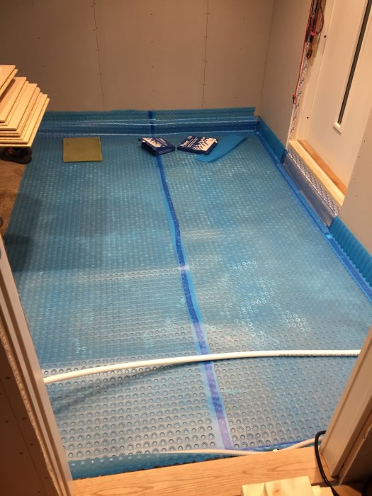 Påbörjad installation av blått golvvärmesystem med bubbelplastlik struktur på betonggolv i ett rums hörn.