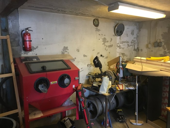 Hörn av ett rörigt garage med betongväggar, diverse verktyg och en röd sandblästrare.