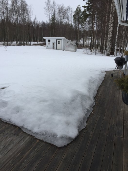 Snötäckt trädgård med stor altan, snösmältning i förgrunden och skogsbakgrund.