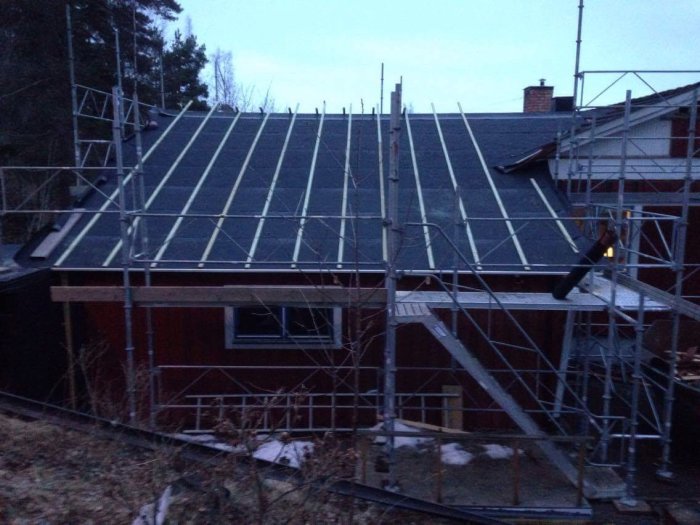 Hus under renovering med byggnadsställningar, snörester, skymning, takpannor, träd i bakgrunden.