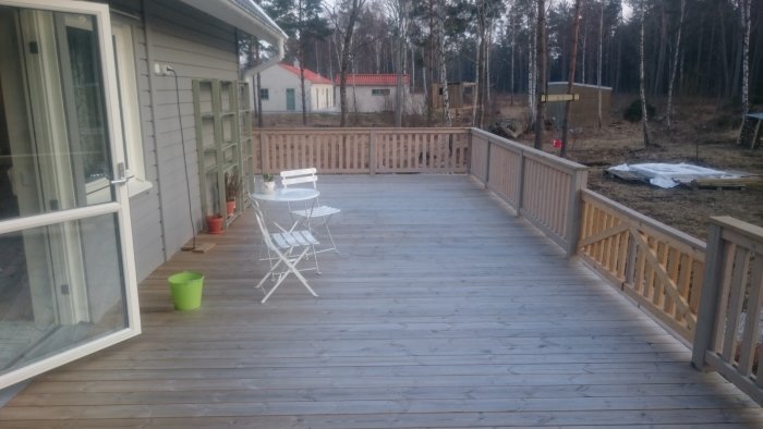 Träterrass vid hus med vitt hopfällbart bord och stolar, passande för utemöblering.