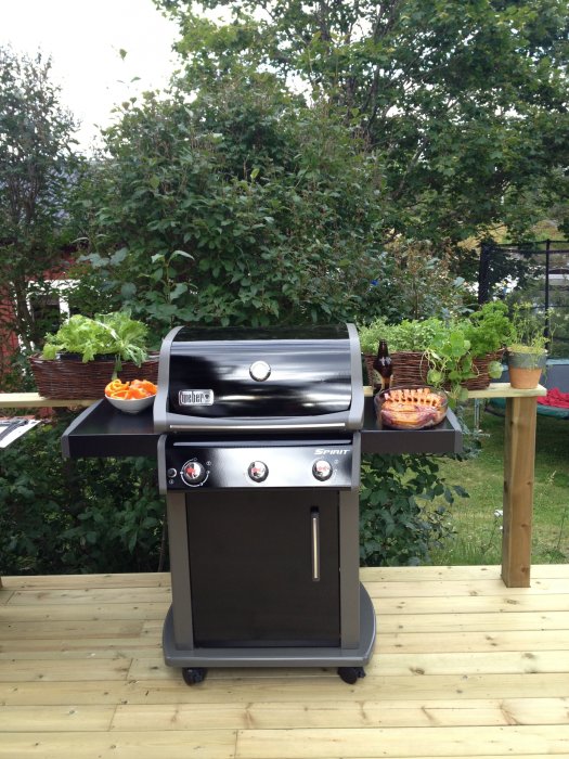 Weber Spirit gasolgrill med tre brännare på en träterrass, omgiven av grönsaker och bladverk.
