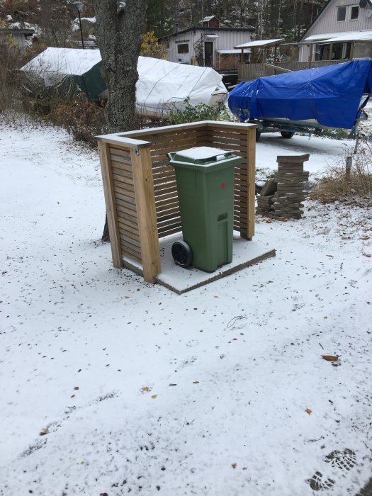 Träskelett som omsluter en grön sophink på en snöig mark, omgiven av träd och båtar täckta med presenningar.