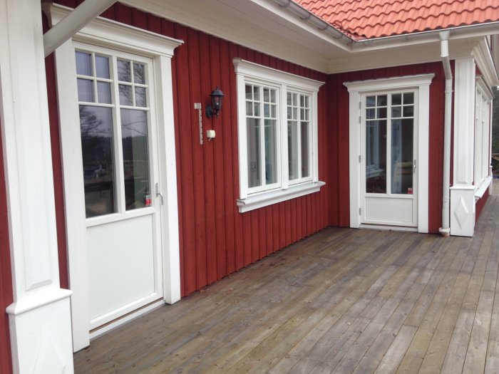 Träveranda utan möbler utanför ett rött hus med vita fönsterkarmar och en ytterdörr, avsett för en framtida sittgrupp.