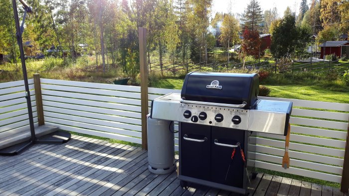 Ett stort gasolgrill på en träaltan med en vit grind framför en naturskön bakgrund av träd och gräsmark.