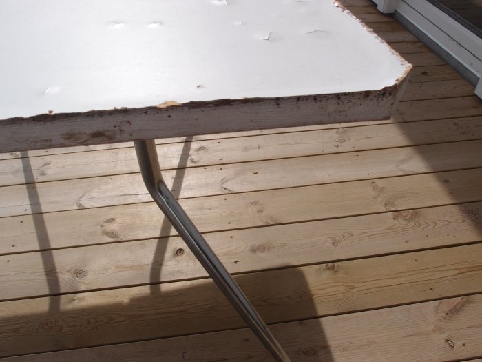 Skadat vitt köksbord med rost och fuktskador på en träaltan i solljus.