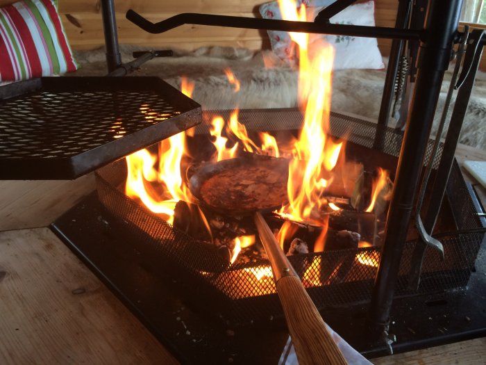 Kolbullar steks över en öppen eld i en grillkåta, med en stekspade i förgrunden och mysig inredning synlig i bakgrunden.