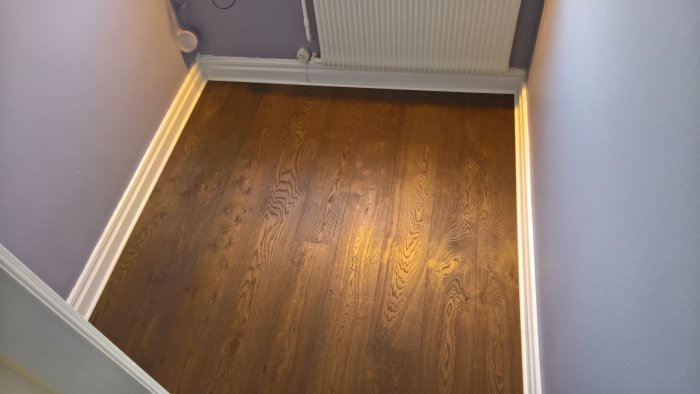 Mörk parkettgolv i ett hörn av ett rum med ljuslila väggar och en vit element, resultat efter renovering.