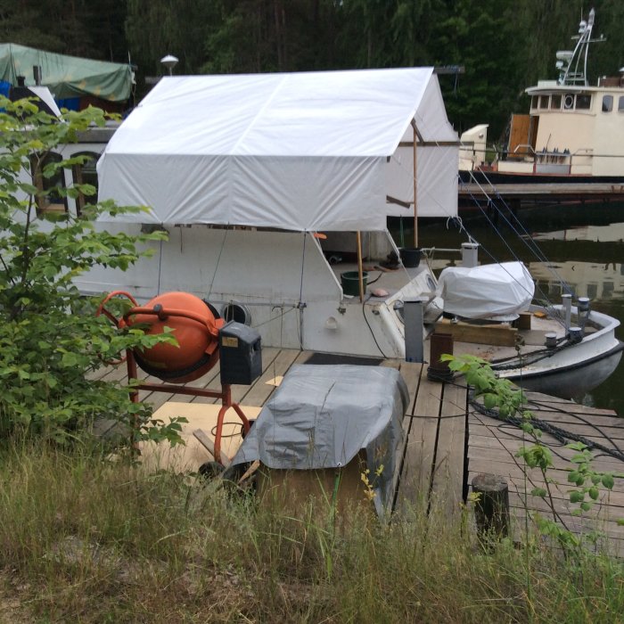 En båt under ett vitt presenningstak vid en brygga med en räddningsboj och en roddbåt i förgrunden.