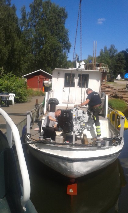 Två personer installerar en ny motor på en båt vid en kaj.