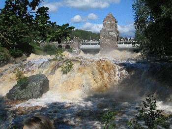 Brusande vattenfall med dimma nära en stenbro och ett torn.