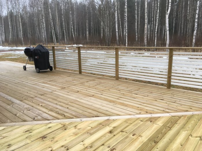 Nybyggd tom uteplats i trä angränsande till skog med en grill till höger och snöfläckar i bakgrunden.