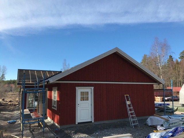 Nybyggd röd stuga med vit ytterdörr, vindskivor och fönsterbläck under klar himmel, byggmaterial synligt.