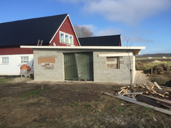 Ofärdigt hus med betonggrund och träbyggnad ovanpå, omgivet av byggmaterial och öppen landsbygd i bakgrunden.