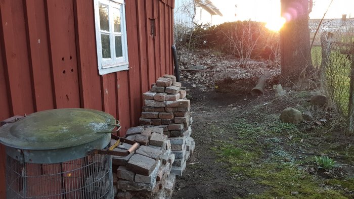 Staplad tegelsten vid en röd trävägg bredvid ett slitet utegrill vid skymning, med en utgrävd plats i bakgrunden.
