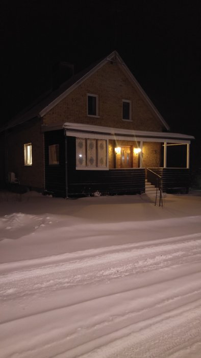 Hus med täckt altan i snölandskap på natten, upplyst av varmt ljus från fönster och dörr.