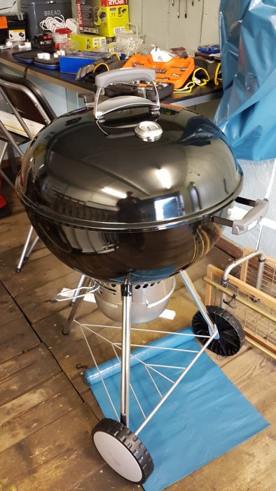 Nybyggd svart Weber 57cm kolgrill står på en blå markskydd i en verkstad med verktyg och lådor i bakgrunden.