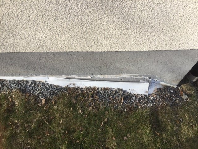 Dräneringslist som har lossnat från husgrund med synlig makadam och gräs i förgrunden.