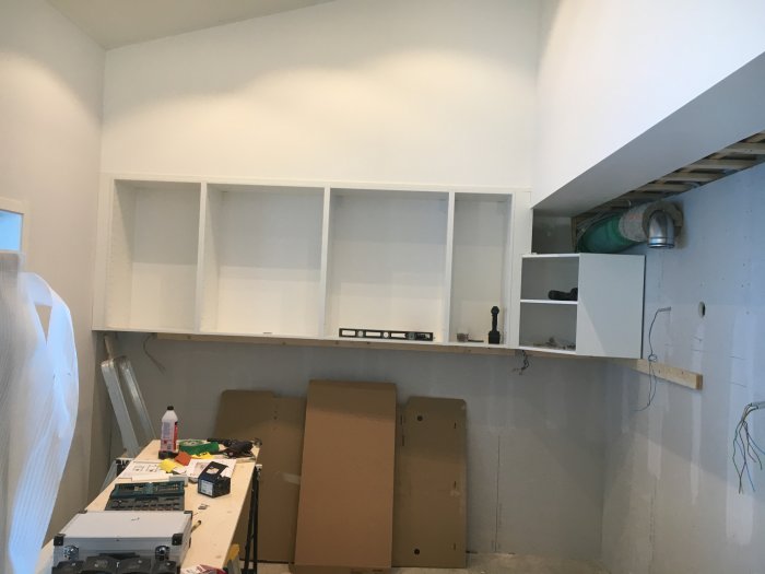 Oinredda köksskåp på vägg, arbetsbänk med verktyg och material, inredningsfas av köksrenovering.