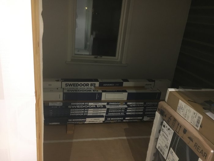 Staplar av oöppnade Swedoor dörrpaket i ett mörkt rum som väntar på installation, samt kartonger.