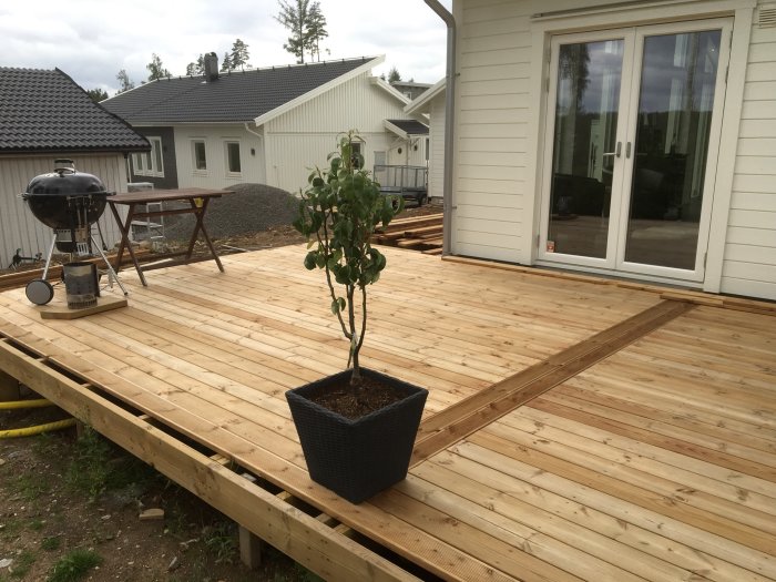 Nybyggd altan med slät brun trall och detalj av rillad trall, omgiven av en vit husfasad och en grill.