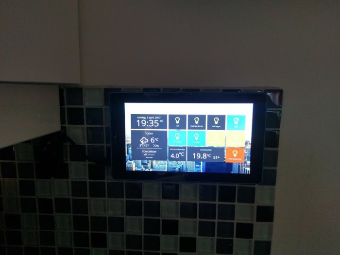Lenovo Tab monterad på vägg som visar tid och väder på hemskärm med färgglada ikoner.