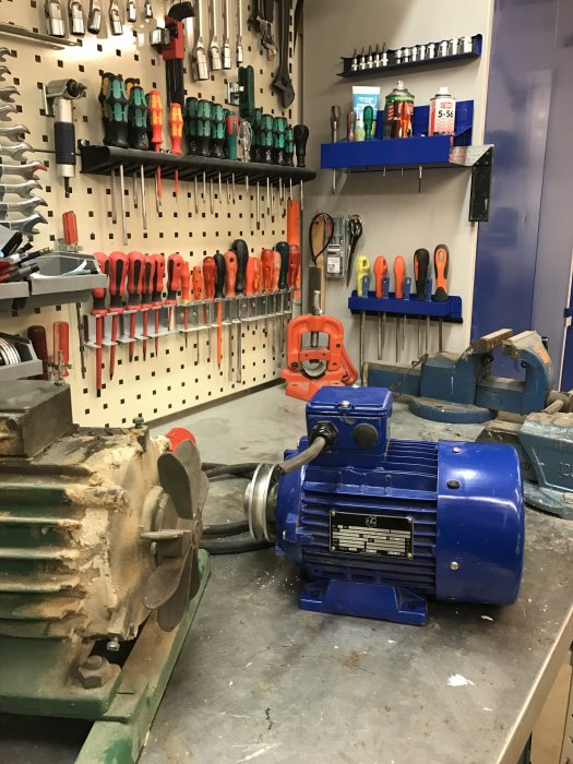 En ny blå elmotor framför en äldre bandsåg i ett verktygsfyllt arbetsrum.