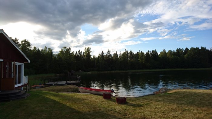 Röd träbåt vid kajkanten nära en brygga vid en sjö med en skog i bakgrunden och del av ett hus.