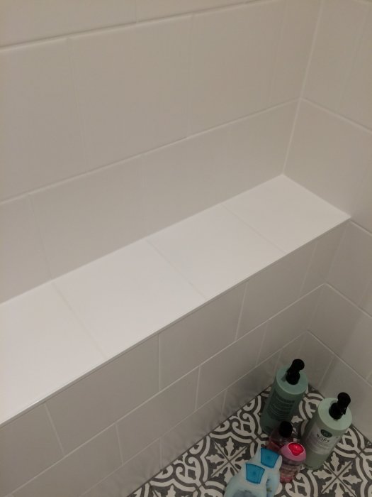 Väggmonterad vit hylla i dusch med kakel och tvåflaskor schampo på golvet.