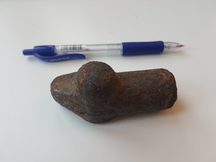 Rostig järndel bredvid en penna ger storleksjämförelse, trolig del från gammal golvbrunn.