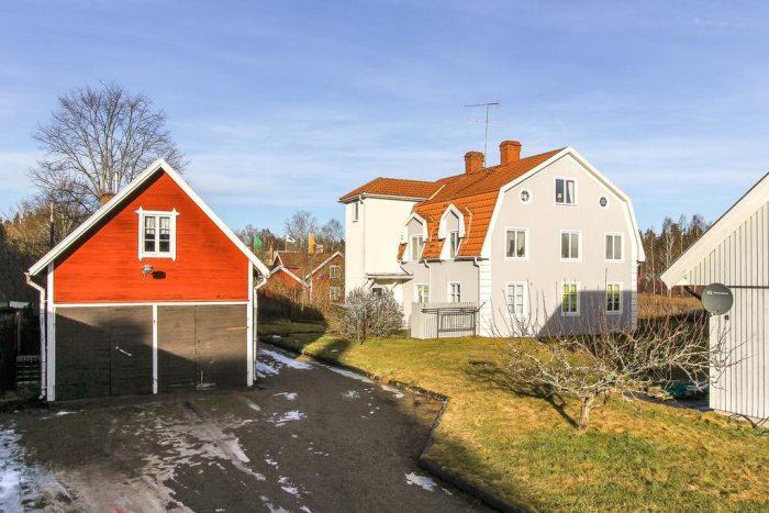 Röd garagelänga bredvid ett större, vitt hus med liknande takvinklar under en klarblå himmel.